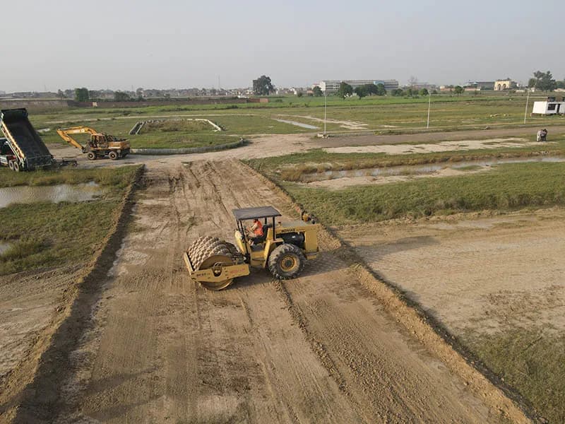 A roller compactor working on a freshly graded road section at the Prism City Lahore site, with an excavator and truck visible nearby.