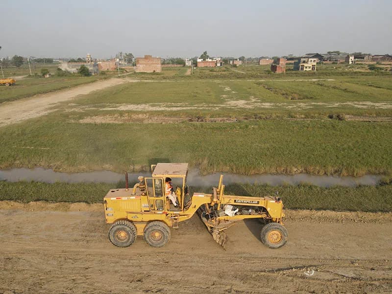 A motor grader levelling ground at the Prism City Lahore development site, with open land and existing structures visible in the background.
