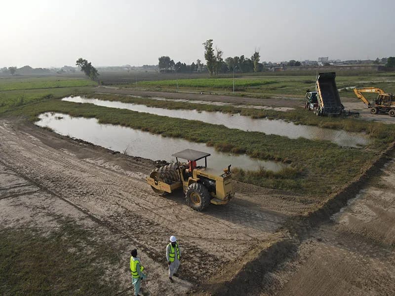 Wider view of ground preparation with a compactor, dump truck, excavator and two workers in high-visibility vests at the Prism City site.